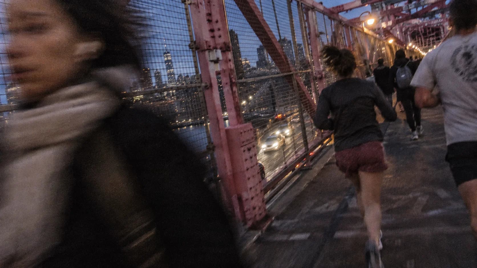 Runners moving across a steel bridge as a blurred passerby crosses the foreground.
