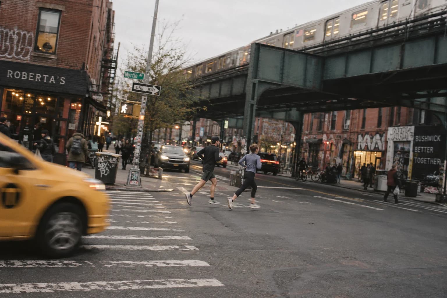 Two runners cross a Brooklyn intersection beneath an elevated train, with a passing taxi blurred in the foreground.